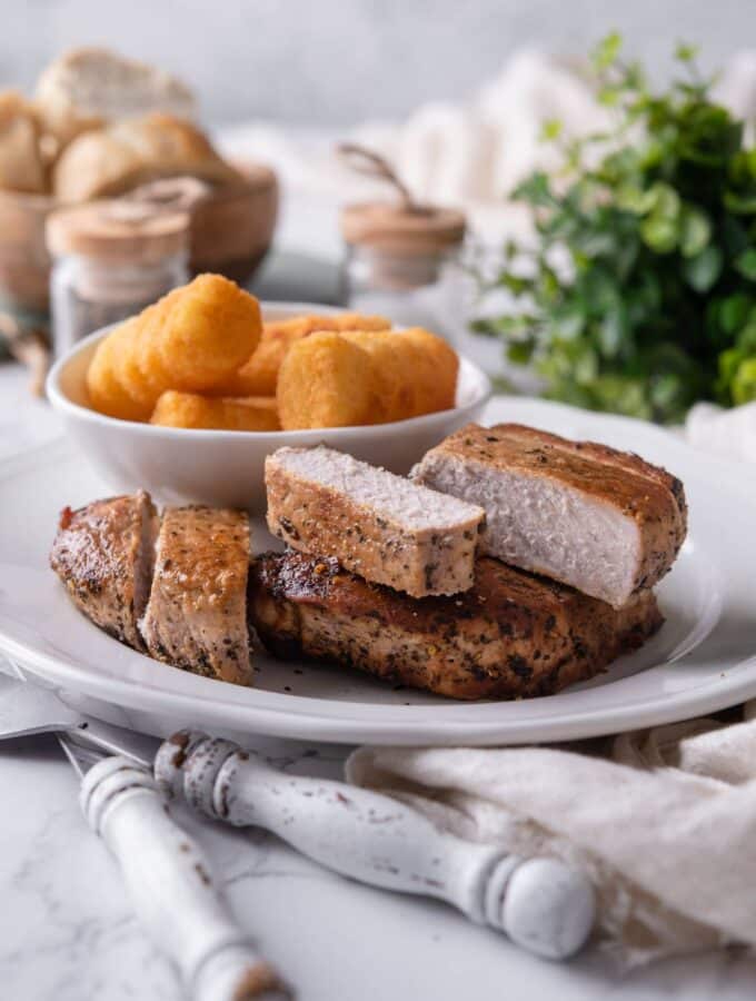 Thick cut pork chops an a white plate with a bowl of tater tots on the side.