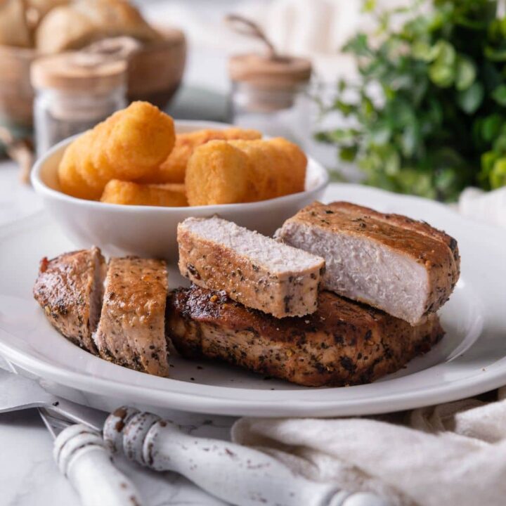 Thick cut pork chops an a white plate with a bowl of tater tots on the side.