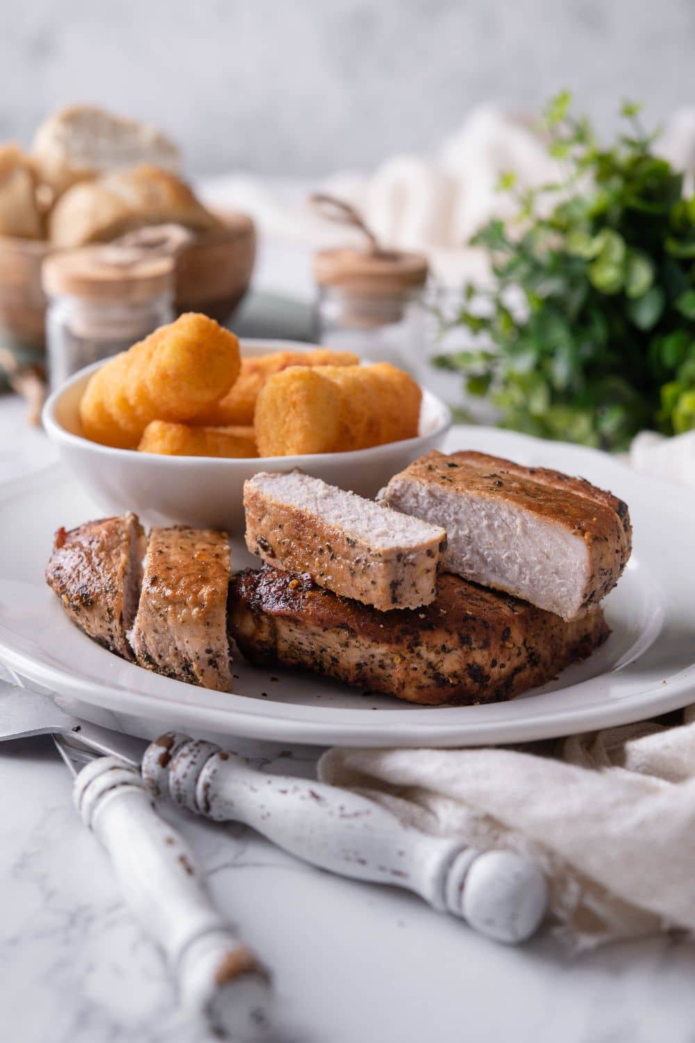 Thick cut pork chops an a white plate with a bowl of tater tots on the side.