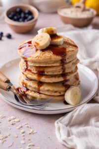 A stack of oat flour pancakes topped with banana slices and drizzled with date syrup on a plate with two forks.