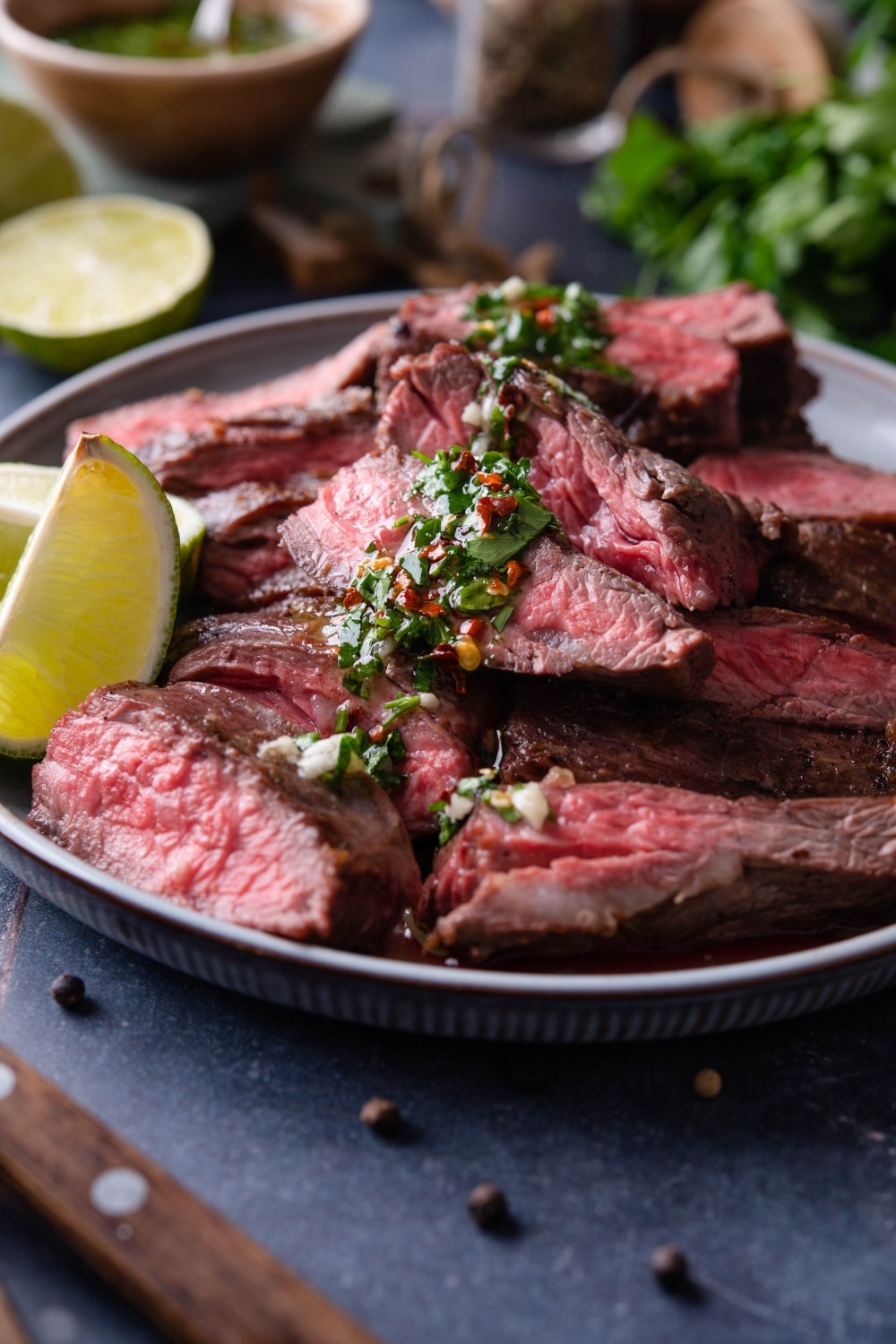 Slices of medium rare skirt steak slices on a plate drizzled with chimichurri and served with lime wedges.