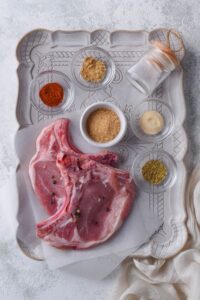 A tray with bone-in pork chops on baking paper alongside small bowls of brown sugar, paprika powder, garlic powder, ginger powder, dried oregano, and a small container of salt.