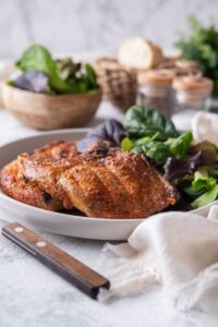 Two broiled bone-in pork chops with salad greens on a plate, with bowls of salad greens and bread slices in the back.