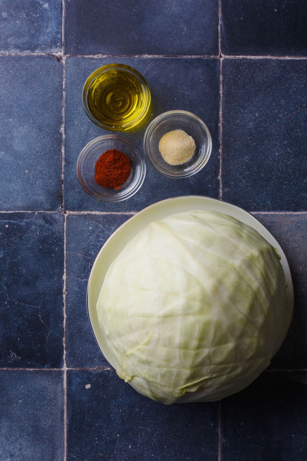 Half a cabbage on a plate next to small bowls of olive oil, garlic powder, and paprika.