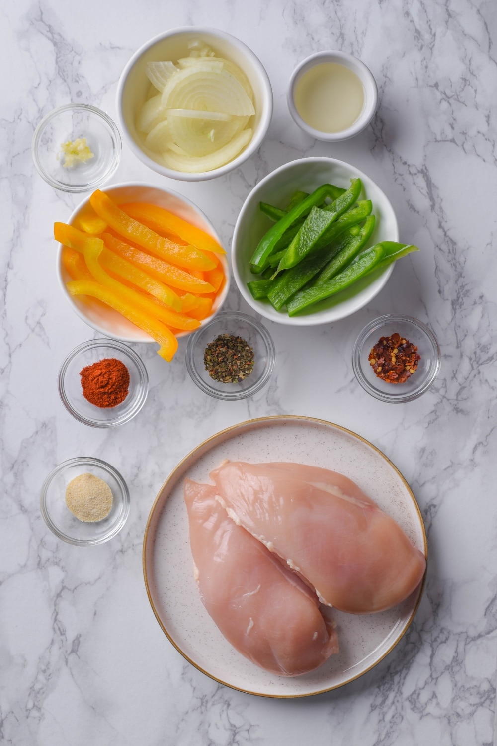 A plate of raw chicken breast fillets, medium-sized bowls of sliced yellow bell peppers, sliced green bell peppers, and sliced onions, and small bowls of individual spices, minced garlic, and oil.