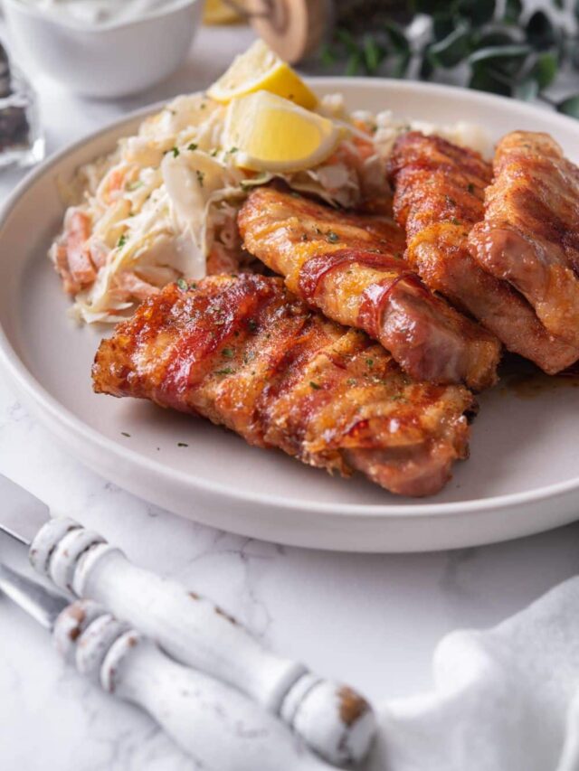 Four bacon wrapped pork chops on a plate with a side of coleslaw and lemon wedges.