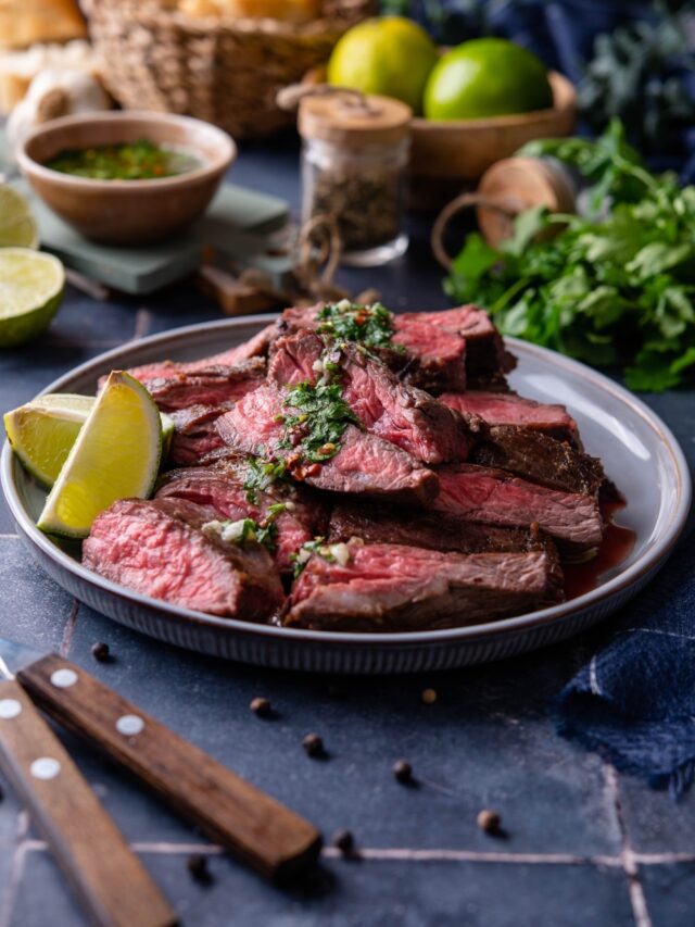 Medium rare skirt steak slices on a plate topped with chimichurri and served with lime wedges. Behind is a bowl of bread, limes, and a small bowl of chimichurri.