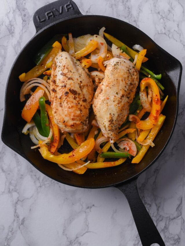 Seared seasoned chicken breasts over sliced bell pepers and onions in a cast iron skillet.