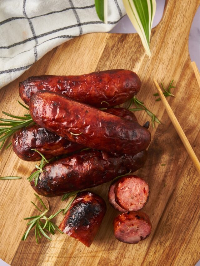 Oven roasted sausages, one sliced into bite-sized pieces, on a wooden serving board. A pair of wooden tongs is resting on the board.
