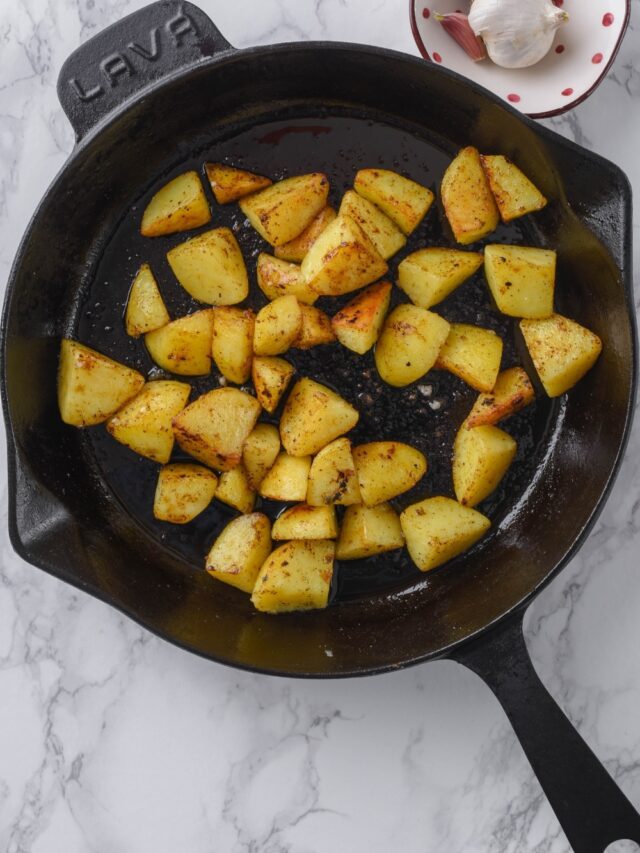 Chopped potatoes sauteeing in a cast iron skillet next to a small bowl of garlic bulbs.