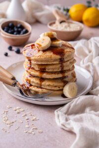 Oat flour pancakes with banana slices and date syrup on a white plate. In the back is a small bowl of blueberries and a bowl of rolled oats.