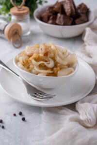 Sauteed onions in a white bowl on a white plate. Behind it is a bowl of steak bites and a pair of salt and pepper shakers.