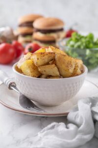 Sauteed potatoes in a white bowl, in front of a bowl of salad and some stacked hamburgers.