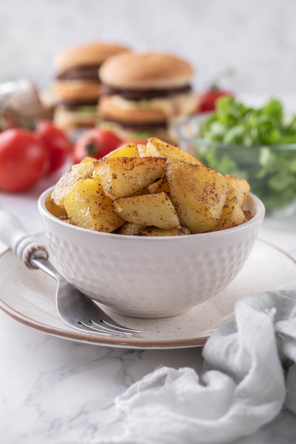 Sauteed potatoes in a white bowl, in front of a bowl of salad and some stacked hamburgers.