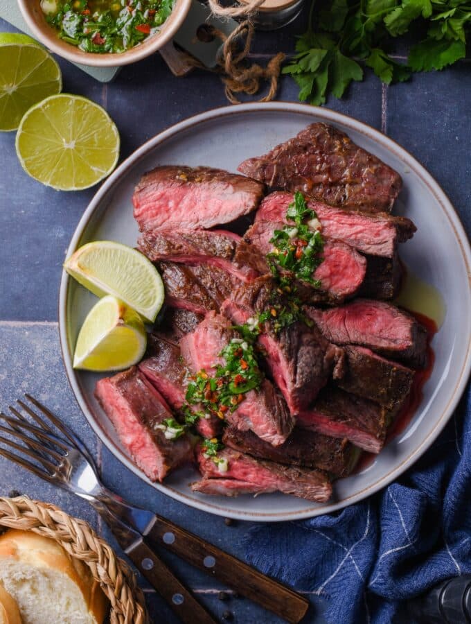 Top view of medium rare skirt steak slices on a plate topped with chimichurri and served with lime wedges. Surrounding it is a small bowl of chimichurri, part of a basket of bread, two forks, fresh cilantro, and a halved lime.