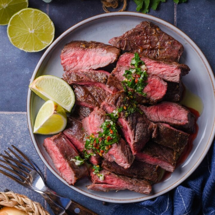 Top view of medium rare skirt steak slices on a plate topped with chimichurri and served with lime wedges. Surrounding it is a small bowl of chimichurri, part of a basket of bread, two forks, fresh cilantro, and a halved lime.