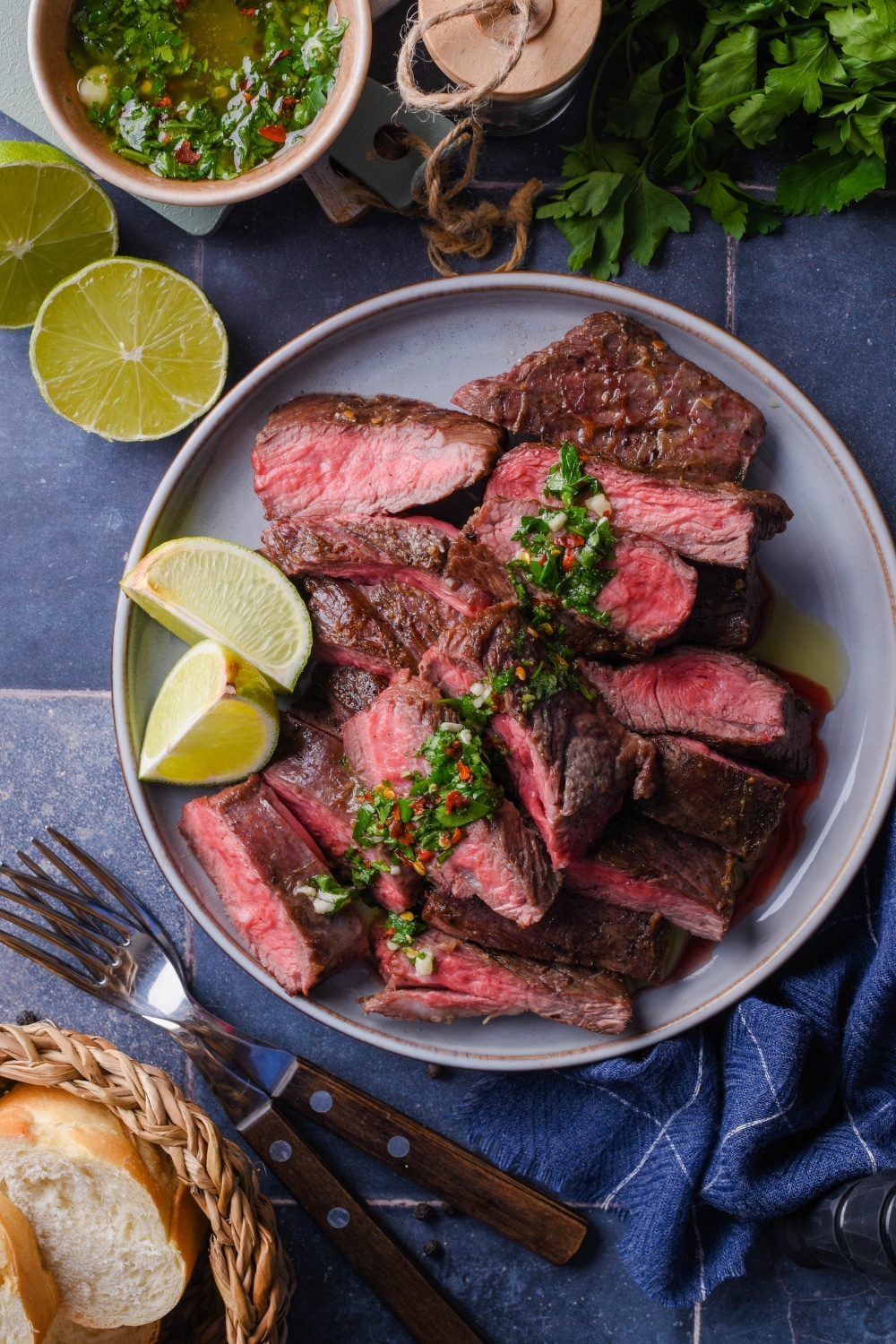 Top view of medium rare skirt steak slices on a plate topped with chimichurri and served with lime wedges. Surrounding it is a small bowl of chimichurri, part of a basket of bread, two forks, fresh cilantro, and a halved lime.