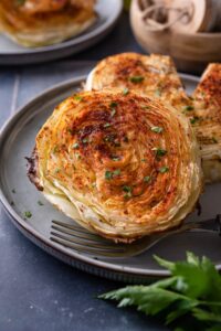 Two roasted cabbage steaks on a plate with a fork, one cabbage steak has a slice taken out.