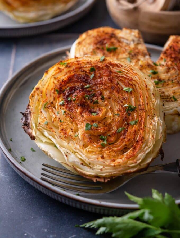 Two roasted cabbage steaks on a plate with a fork, one cabbage steak has a slice taken out.