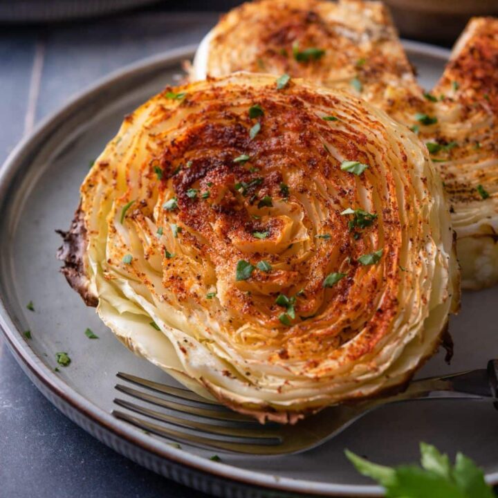 Two roasted cabbage steaks on a plate with a fork, one cabbage steak has a slice taken out.