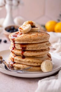 Oat flour pancakes on a plate, drizzled with date syrup and topped with banana slices, served with two forks.