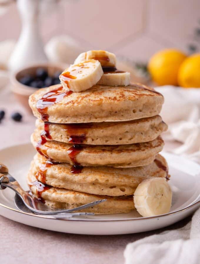 Oat flour pancakes on a plate, drizzled with date syrup and topped with banana slices, served with two forks.