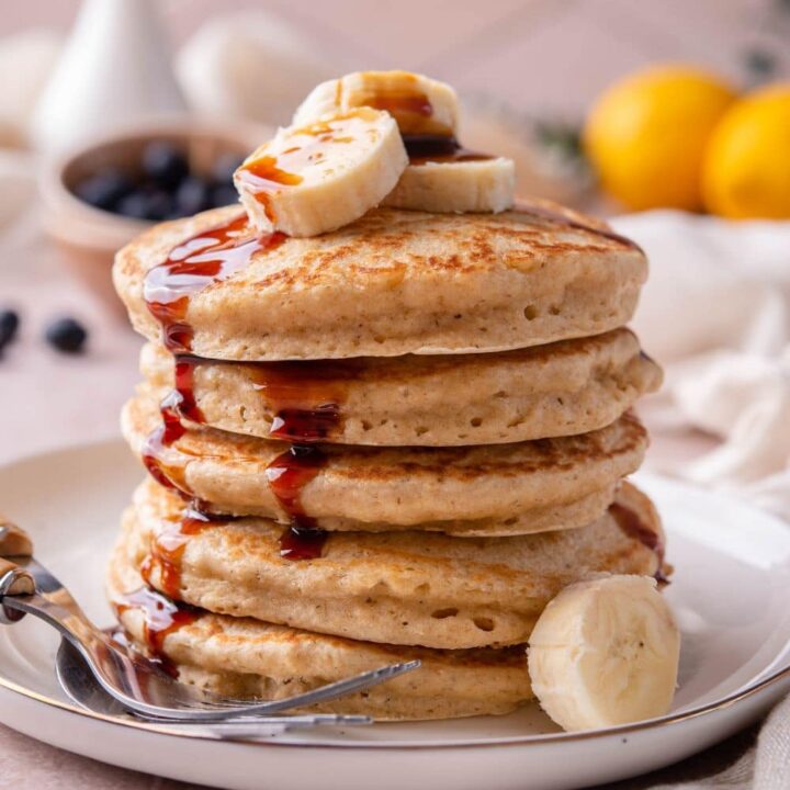 Oat flour pancakes on a plate, drizzled with date syrup and topped with banana slices, served with two forks.