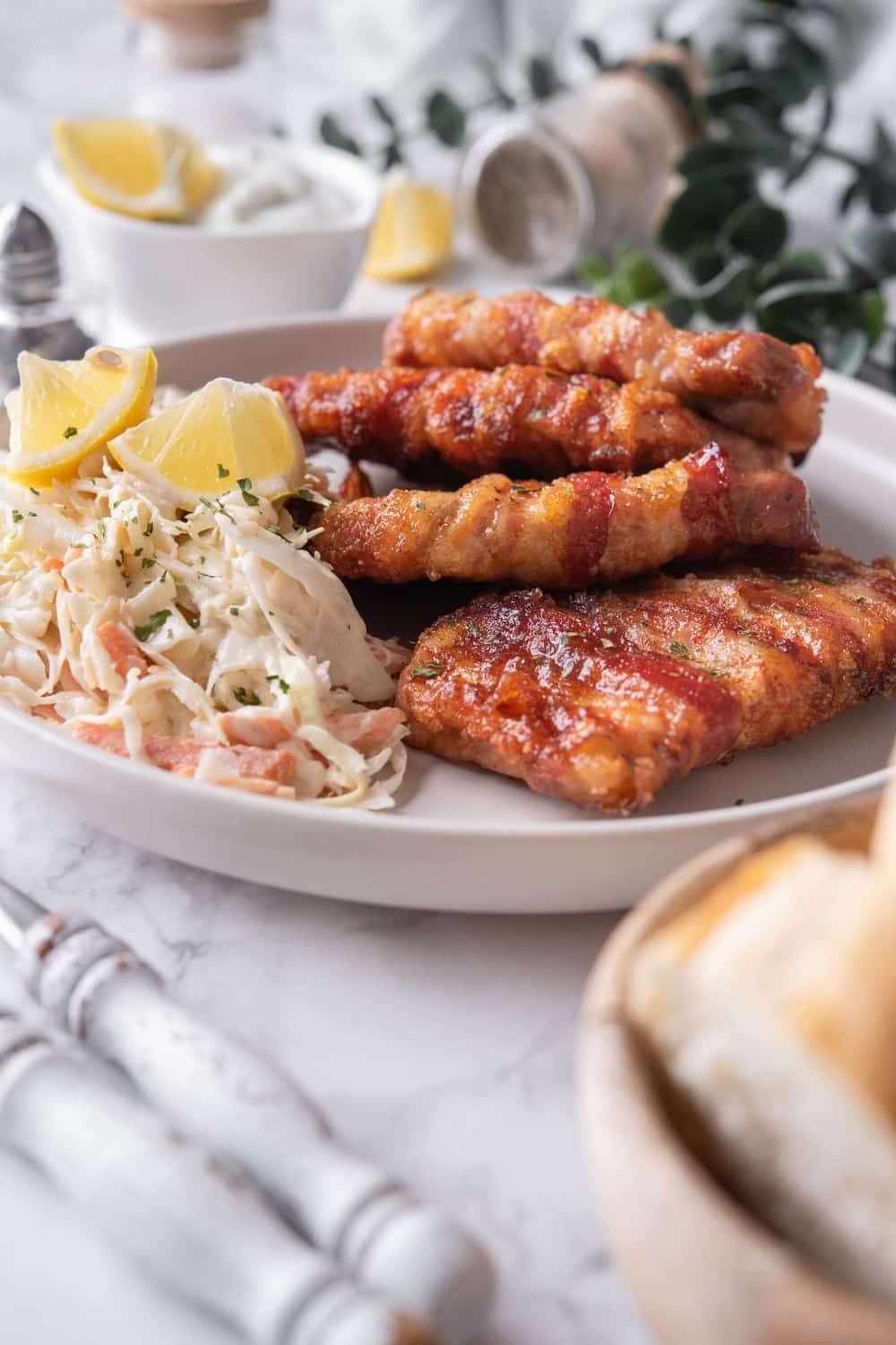 Four bacon wrapped pork chops on a plate with a side of coleslaw with lemon wedges on top, served with a small bowl of creamy dip and a bowl of bread.