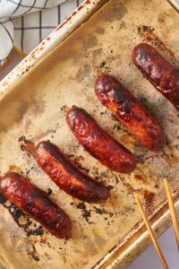 Oven roasted sausages on a parchment paper-lined baking sheet with a pair of wooden tongs.