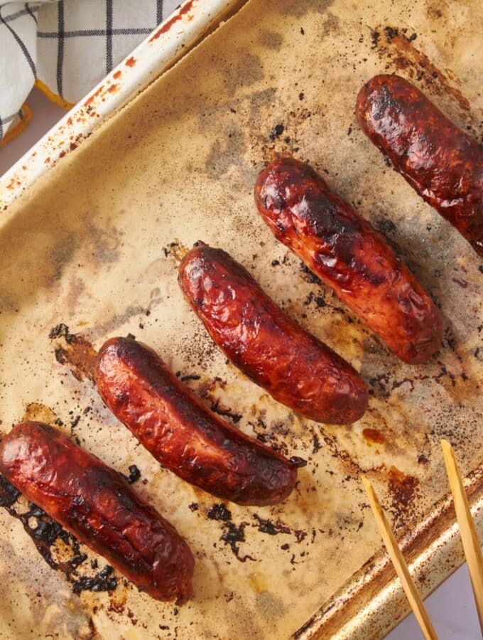 Oven roasted sausages on a parchment paper-lined baking sheet with a pair of wooden tongs.