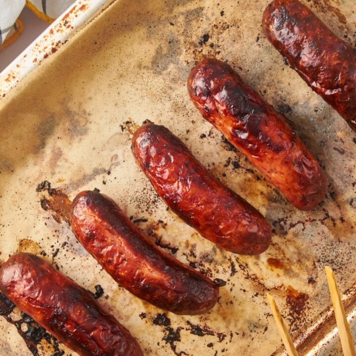 Oven roasted sausages on a parchment paper-lined baking sheet with a pair of wooden tongs.