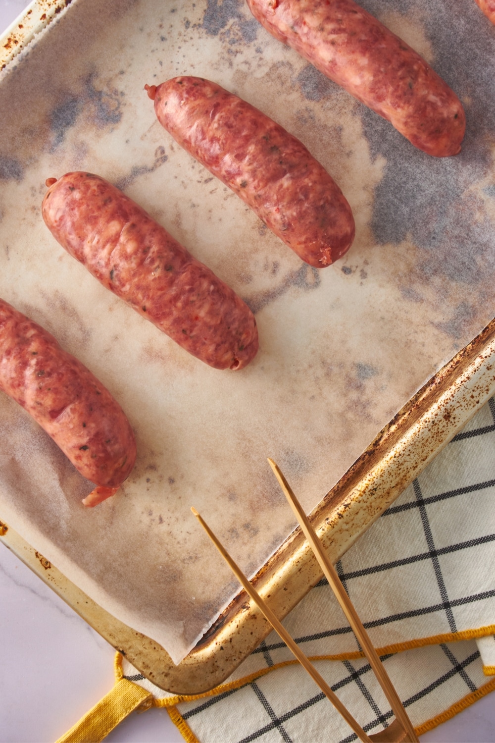 Raw sausages on a parchment paper-lined baking sheet with a pair of wooden tongs.