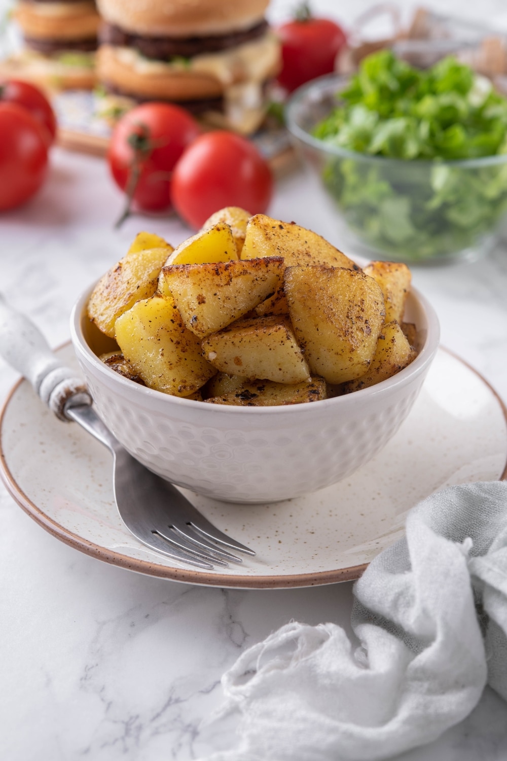 Sauteed chopped potatoes in a white bowl, in front of a bowl of salad and some stacked hamburgers.