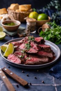 Medium rare skirt steak slices on a plate topped with chimichurri and served with lime wedges. Behind is a bowl of bread, limes, and a small bowl of chimichurri.
