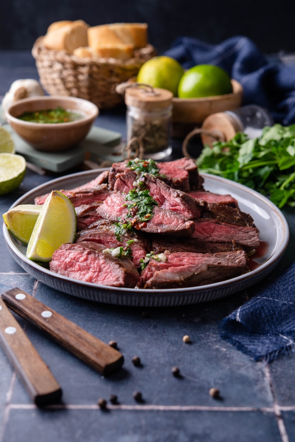 Medium rare skirt steak slices on a plate garnished with chimichurri and lime wedges. Behind are more limes and a small bowl of chimichurri.