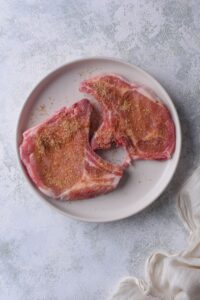 Two uncooked bone-in pork chops coated in spice rub on a plate.