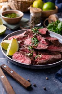 Skirt steak slices on a plate garnished with chimichurri and lime wedges.