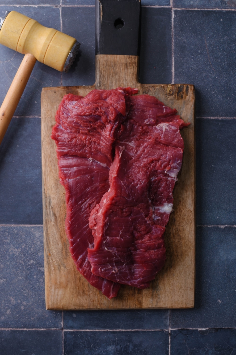 Raw skirt steak on a wooden cutting board next to a meat mallet.