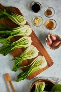 Six bok choy halves on a wooden cutting board. Next to it is a bowl of shallots, a bowl of sesame oil, a bowl of soy sauce, a bowl of garlic, and a bowl of soy sauce.