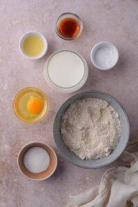 A bowl of oat flour and small bowls of baking powder, raw egg, sugar, milk, and lemon juice.