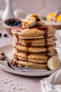 A tall stack of oat flour pancakes on a plate topped with fresh banana slices and drizzled with date syrup.