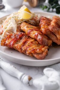 Four bacon wrapped pork chops with a brown sugar and chili rub on a plate with a side of slaw and lemon wedges.