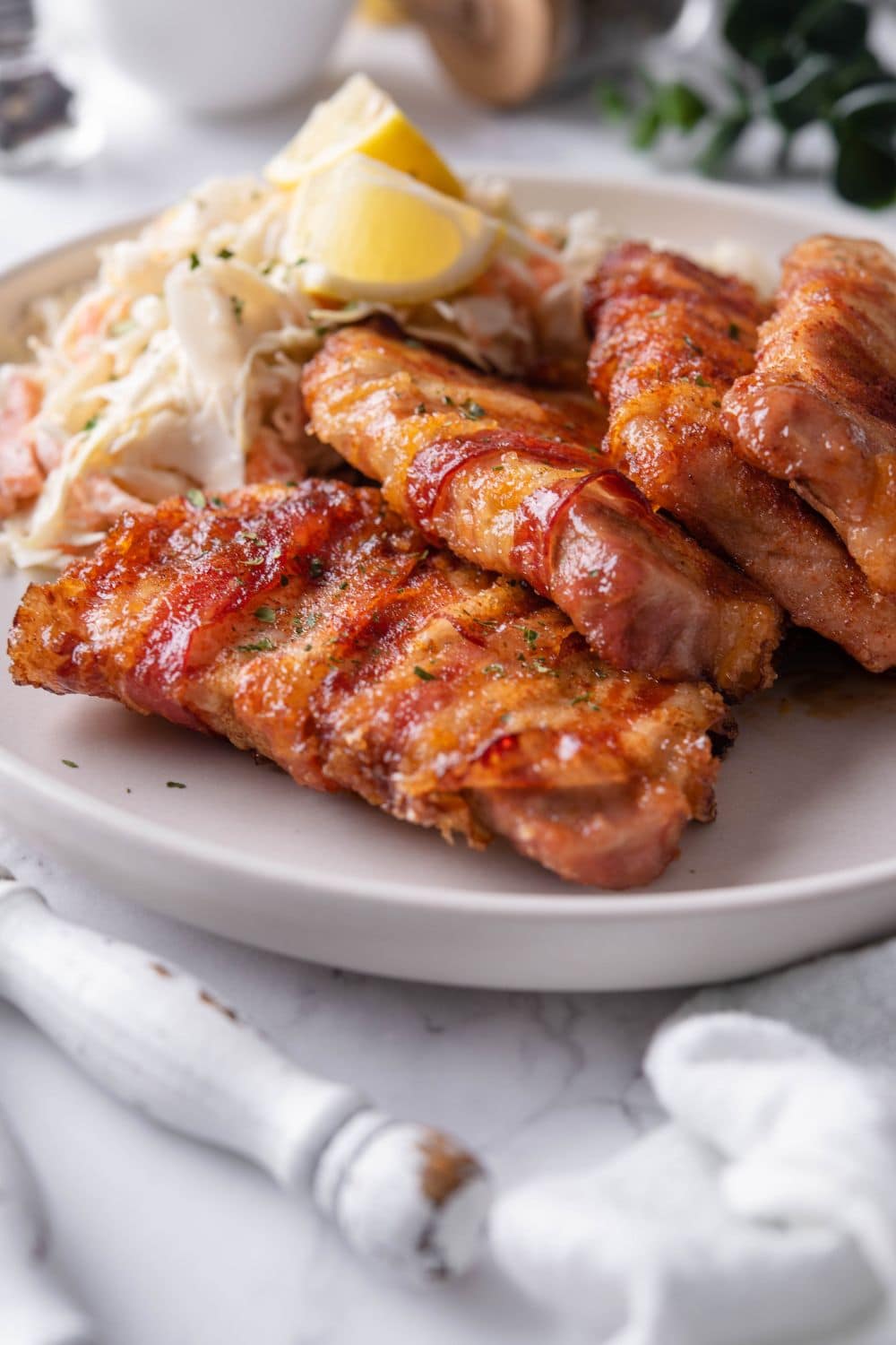 Four bacon wrapped pork chops with a brown sugar and chili rub on a plate with a side of slaw and lemon wedges.