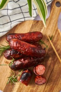 Oven roasted sausages, one sliced into bite-sized pieces, on a wooden serving board. A pair of wooden tongs is resting on the board.