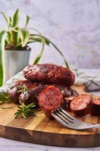 A piece of sliced cooked sausage on a fork. It's resting on a wooden serving board with oven roasted sausages.