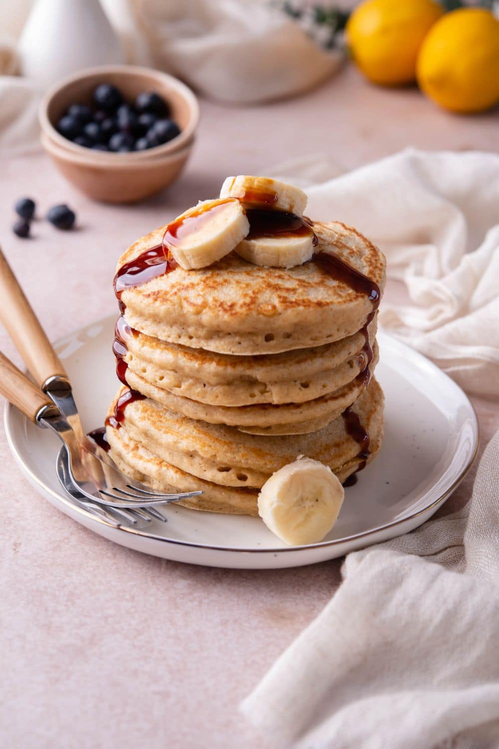 Oat flour pancakes topped with banana slices and date syrup on a plate with two forks and a small bowl of blueberries in the back.