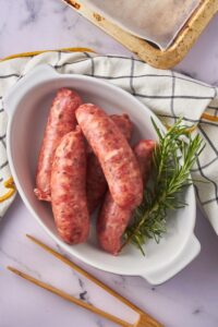 Raw sausages and fresh rosemary in a white oval baking dish. Part of a baking sheet and a pair of wooden tongs surround it.