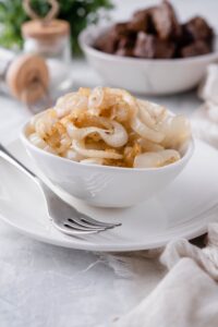 Sauteed onions in a white plate on a white plate, in front of a bowl of steak bites and a pair of salt and pepper shakers.