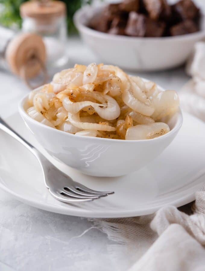 Sauteed onions in a white plate on a white plate, in front of a bowl of steak bites and a pair of salt and pepper shakers.