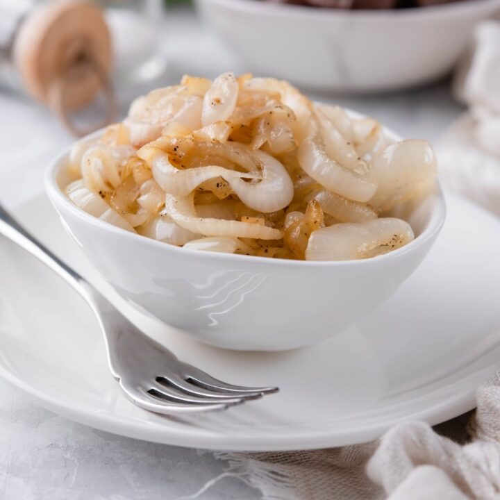 Sauteed onions in a white plate on a white plate, in front of a bowl of steak bites and a pair of salt and pepper shakers.